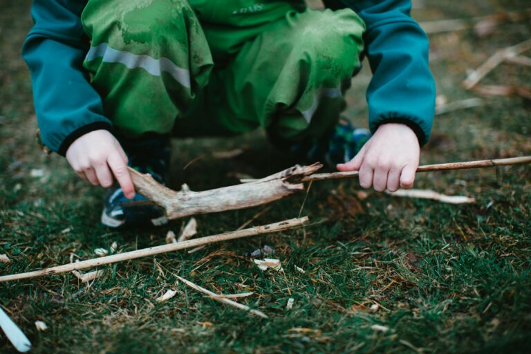 From above of crop faceless squatting child in warm clothes playing with tree branches in forest