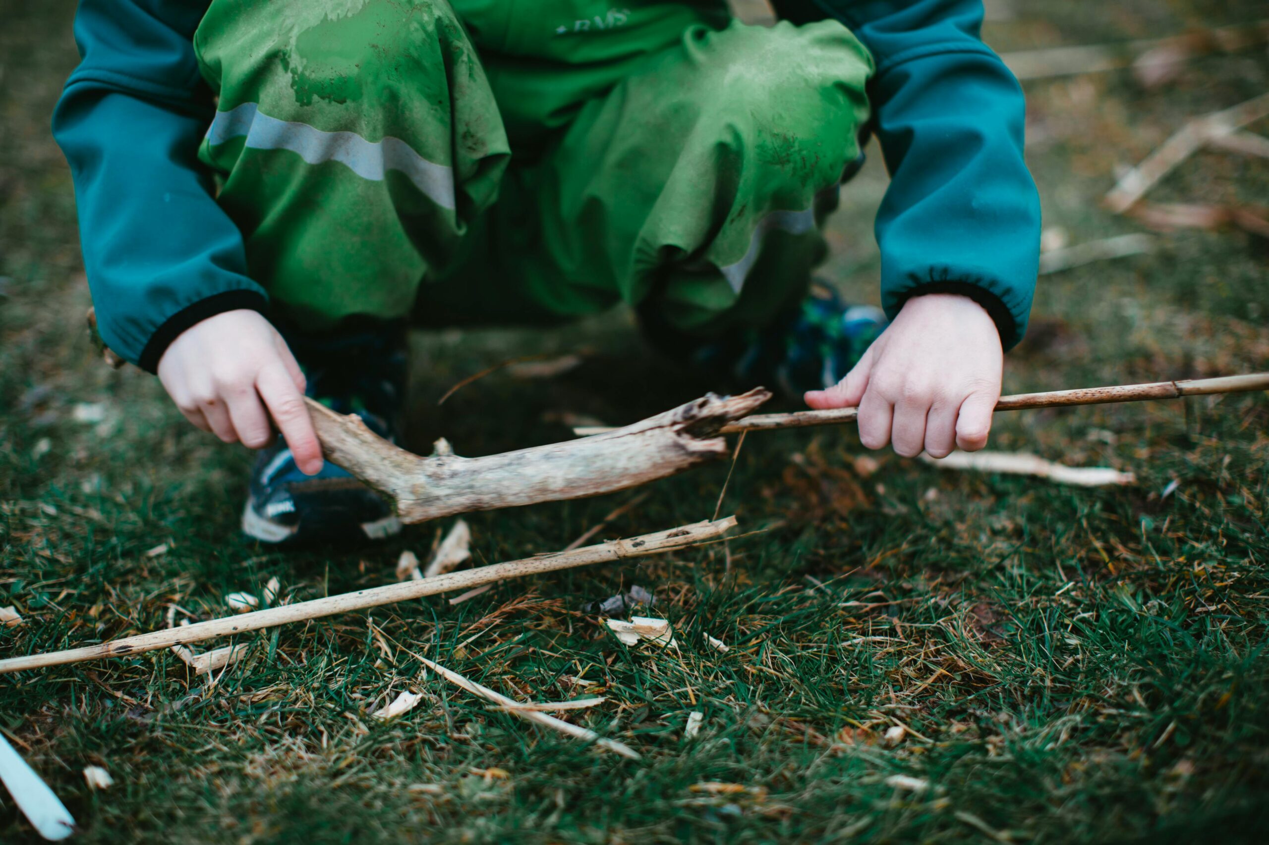 From above of crop faceless squatting child in warm clothes playing with tree branches in forest