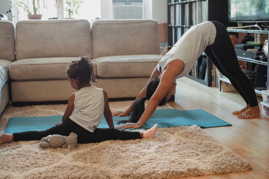 Unrecognizable female and little girl in sportswear stretching on carpet and stretch mat in living room