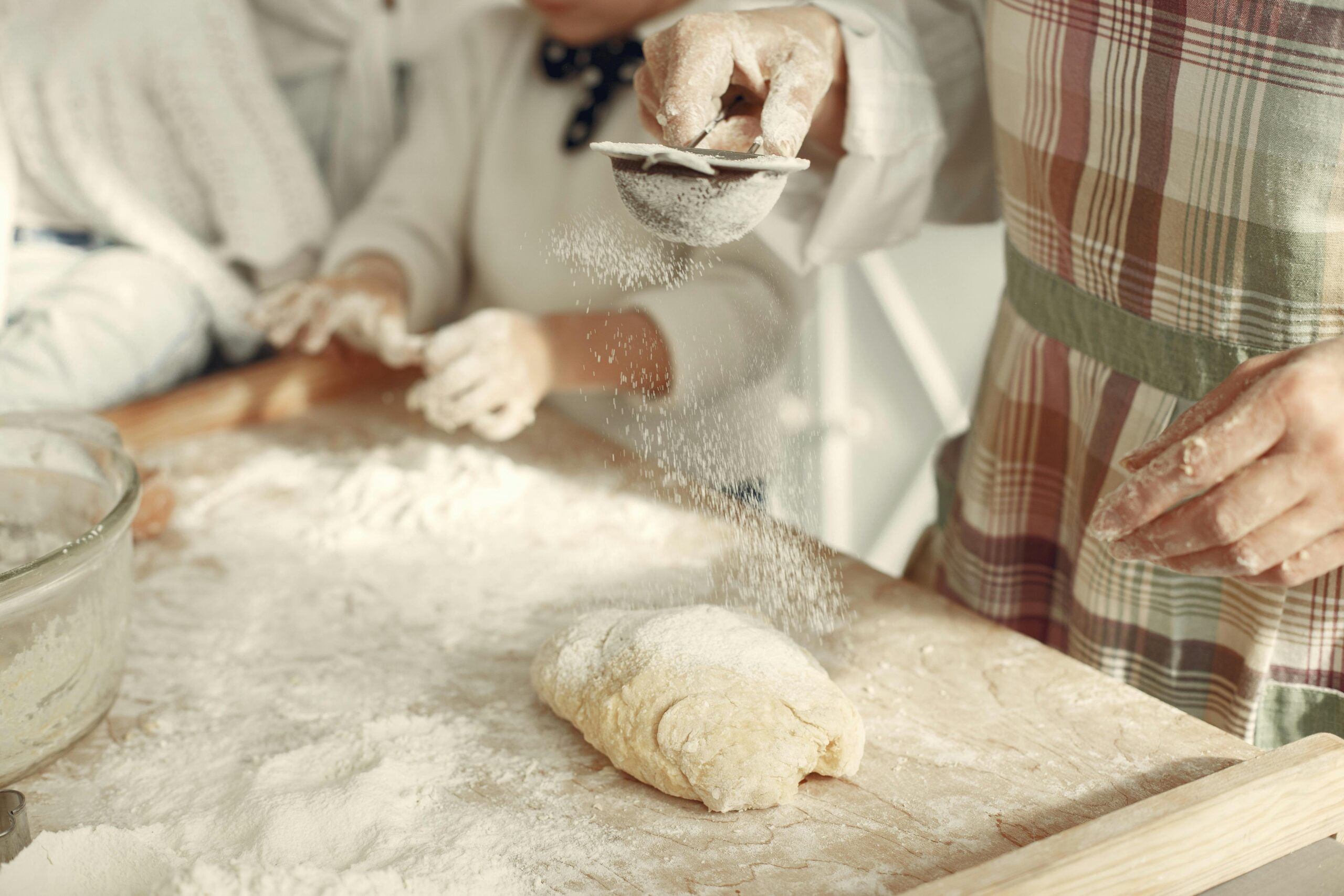 A grandmother and granddaughter baking together, sifting flour onto dough in a warm kitchen setting.