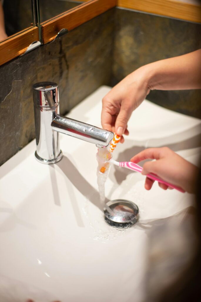 Close-up of hands washing toothbrushes under running faucet in a bathroom setting.