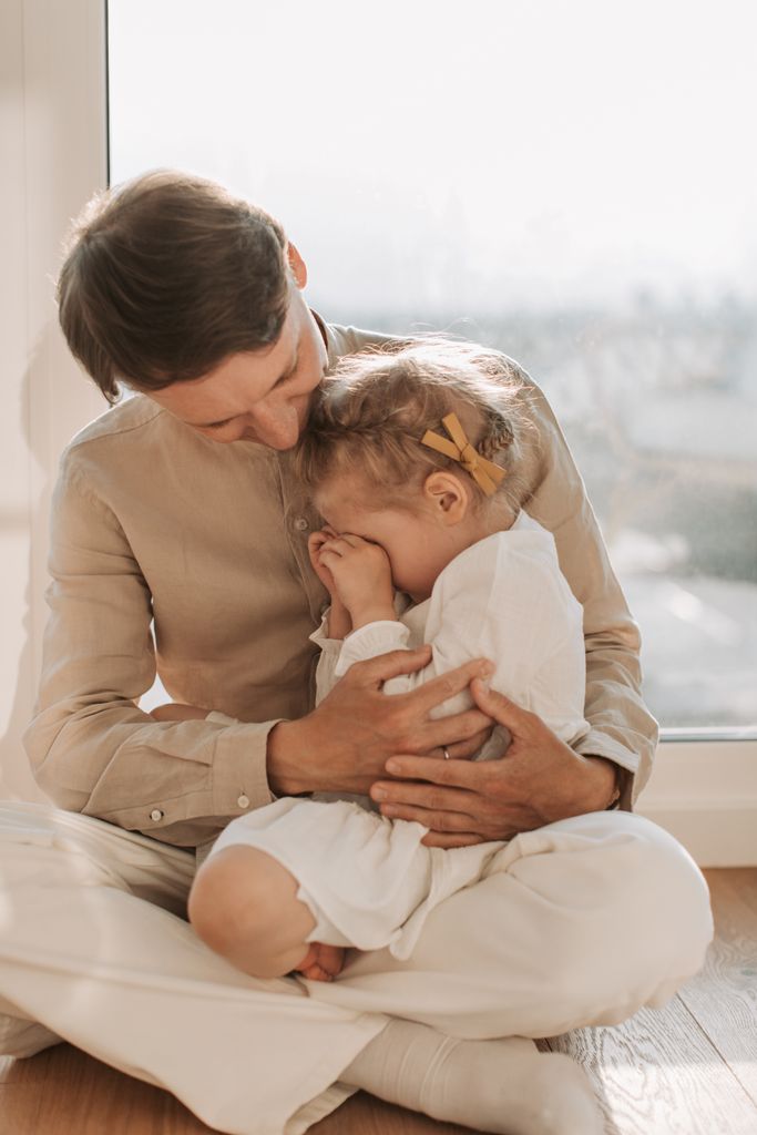 A tender moment between a father and daughter sitting by the window, filled with love and comfort.