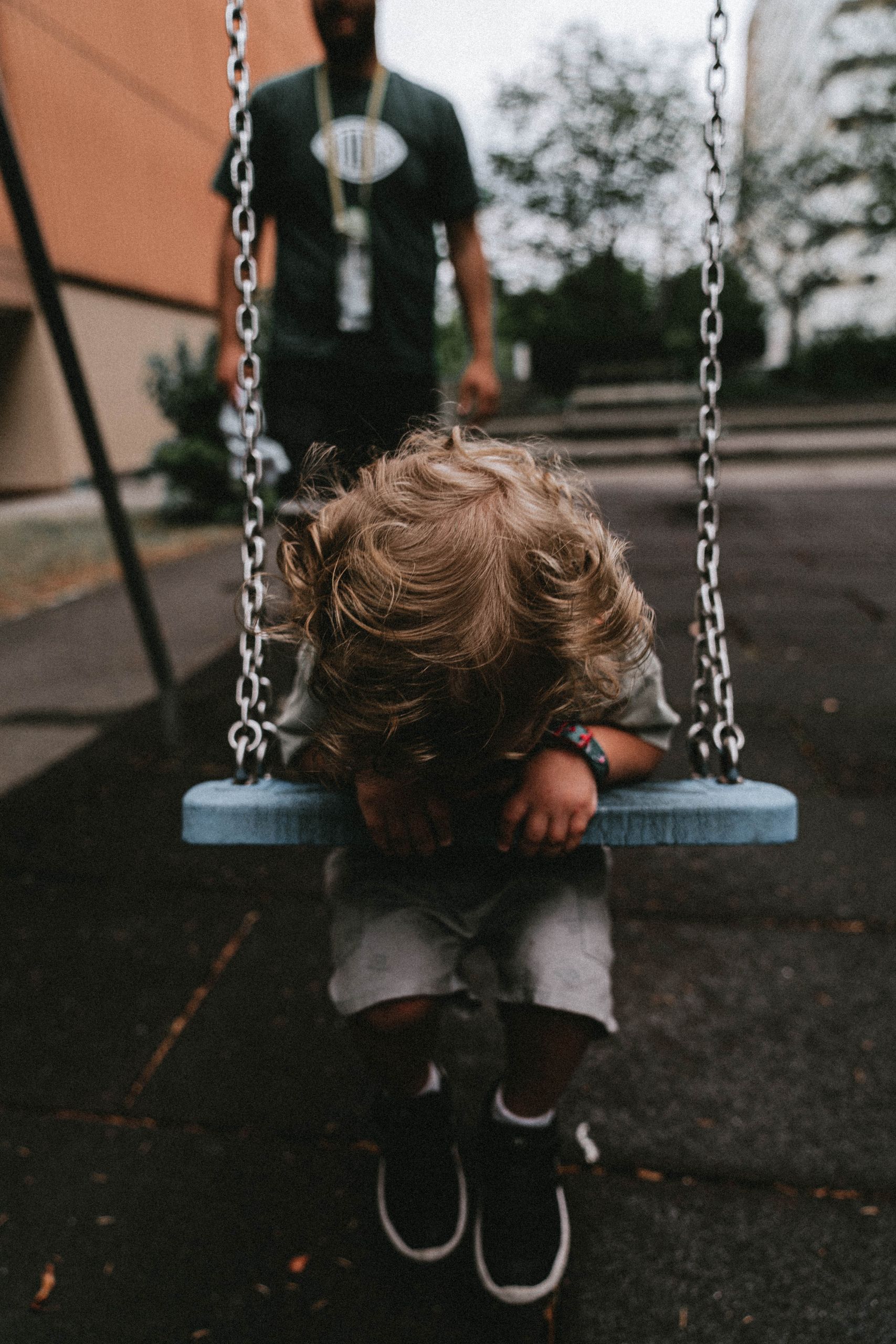 Adorable blond child sitting on a swing with a parent in the background, in a park setting.